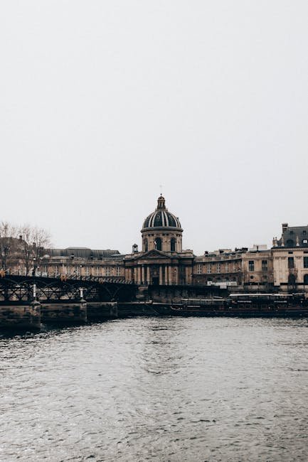 Aged institute facade with columns against bridge above Seine River under light sky in Paris France